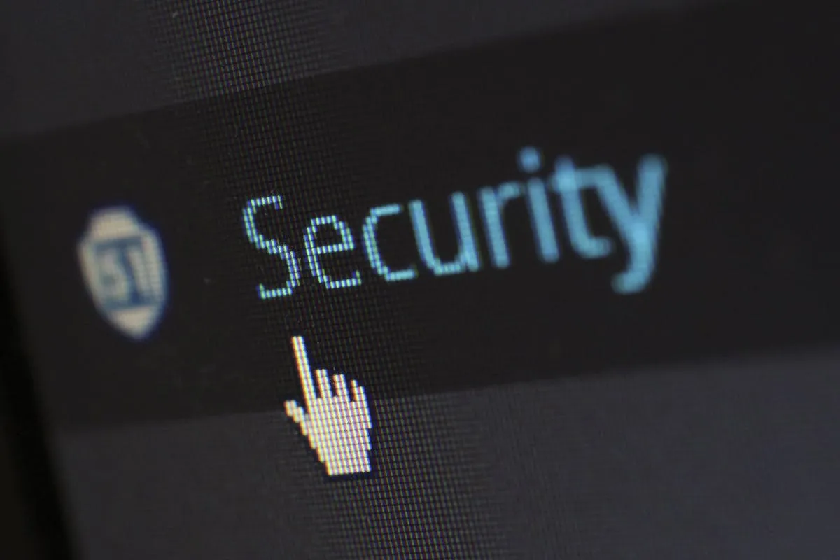 Macro aproximado de uma tela LCD exibindo a palavra Security escrita em azul-claro ao lado de um pequeno ícone de escudo desfocado, com o cursor do mouse em formato de mão apontando para o texto sobre fundo escuro.