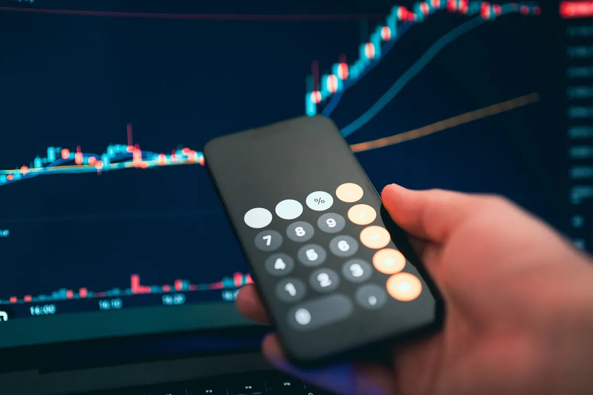 Close-up de uma mão humana segurando um smartphone com o aplicativo de calculadora aberto, em frente à tela de um notebook que exibe um gráfico financeiro de linhas e barras com fundo escuro.