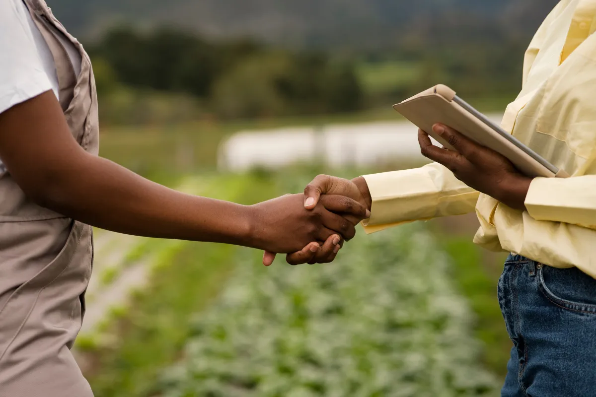Profissionais apertam as mãos em área agrícola, simbolizando acordos comerciais na cadeia de distribuição do agro.