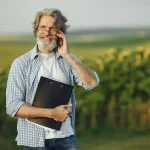 Homem idoso de pele clara, cabelos grisalhos e barba branca, vestindo camisa xadrez azul sobre camiseta branca e calça jeans, sorri enquanto fala ao celular e segura uma prancheta preta com papéis em uma plantação ao ar livre.