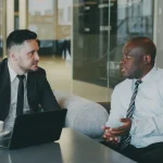 Dois executivos de terno conversam sentados à mesa de uma cafeteria; o homem da esquerda, de pele clara e terno preto, anota em um bloco ao lado de um notebook, enquanto o homem da direita, de pele negra e camisa social clara, fala gesticulando.