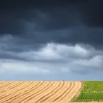 Fotografia em plano aberto e ângulo normal mostrando uma paisagem rural dividida. Na metade inferior esquerda, um campo arado exibe linhas paralelas e curvas de nível em tom de palha claro. Na metade inferior direita, estende-se uma plantação verde e alinhada com uma árvore solitária na linha do horizonte. A metade superior da imagem é inteiramente preenchida por nuvens de tempestade densas, pesadas e escuras em tons de cinza e preto, gerando um contraste dramático com a terra iluminada.