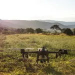 Um drone agrícola de grande porte pousado em meio a um campo verdejante e aberto, com montanhas e árvores ao fundo sob a luz do sol nascente ou poente.
