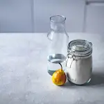 Mão segurando bicarbonato de sódio com produtos de limpeza ao fundo
