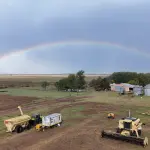 Vista aérea oblíqua de uma propriedade rural com maquinário agrícola em primeiro plano, instalações e silos ao fundo, sob um céu nublado cortado por um arco-íris completo.