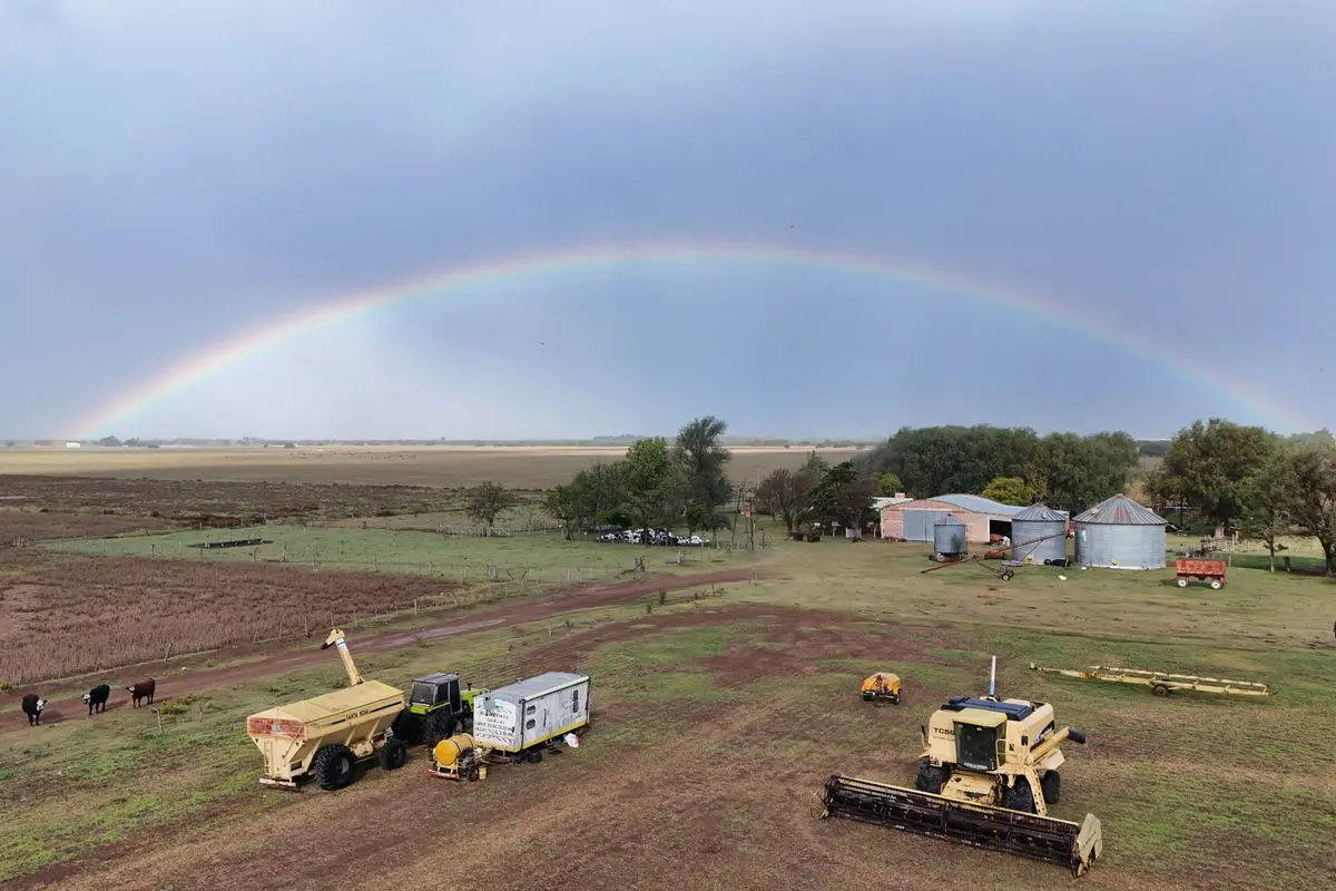 Vista aérea oblíqua de uma propriedade rural com maquinário agrícola em primeiro plano, instalações e silos ao fundo, sob um céu nublado cortado por um arco-íris completo.