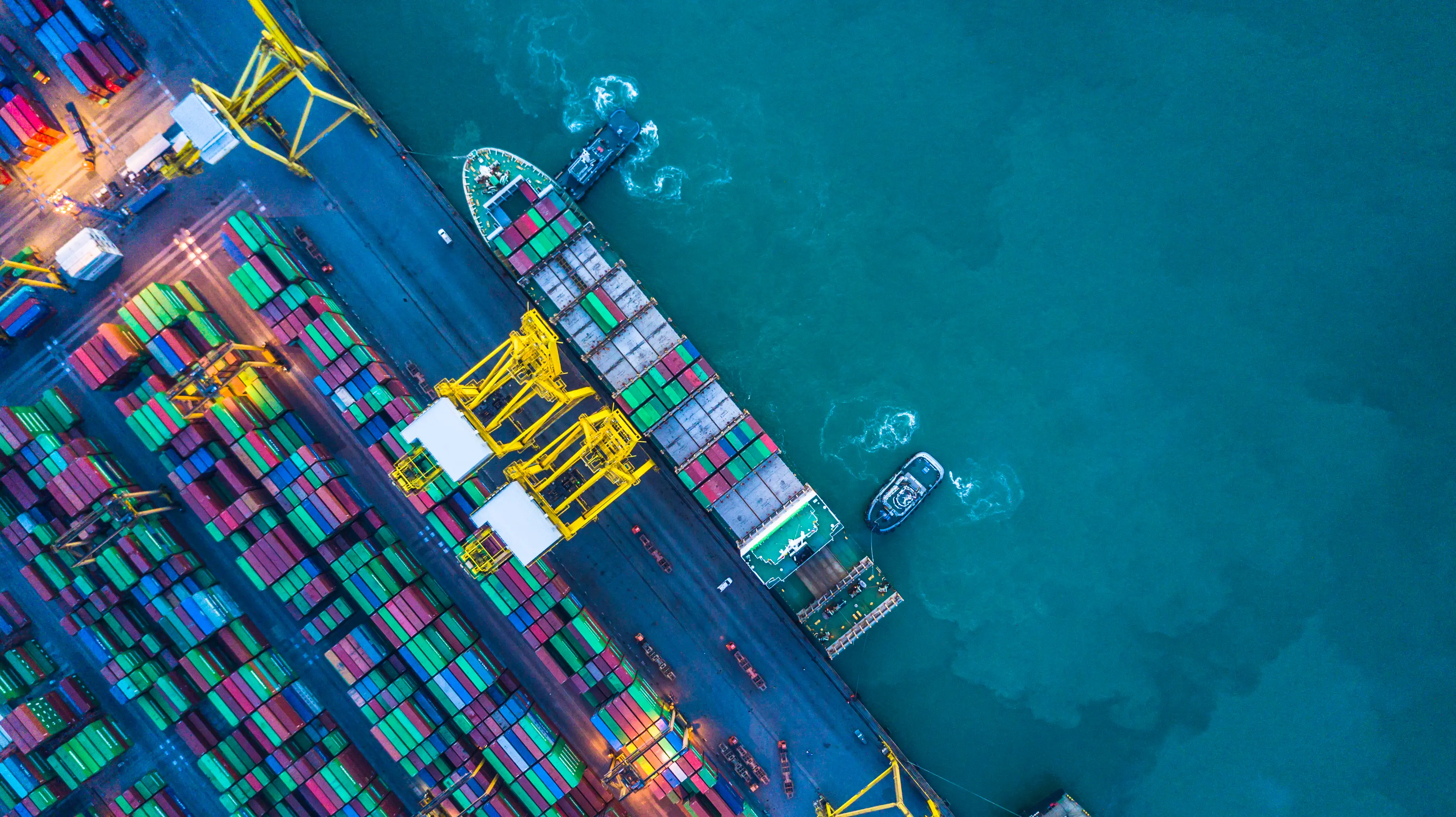 Vista aérea vertical (top-down) de um porto comercial movimentado, mostrando um grande navio cargueiro atracado, dezenas de contêineres coloridos organizados e guindastes amarelos de carga sobre as águas esverdeadas do oceano.