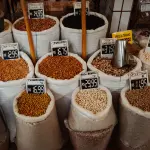 Vista frontal e superior de vários sacos grandes de ráfia e plástico cheios de diferentes tipos de feijões e grãos expostos à venda, cada um com uma placa de preço manuscrita à frente.