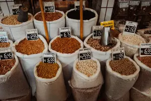 Vista frontal e superior de vários sacos grandes de ráfia e plástico cheios de diferentes tipos de feijões e grãos expostos à venda, cada um com uma placa de preço manuscrita à frente.