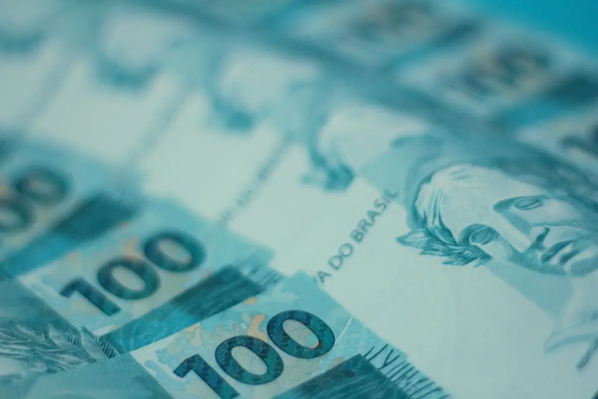 Vista em plano detalhado e inclinado de várias cédulas de cem reais brasileiros dispostas em camadas com foco seletivo no número cem e na efígie da República.