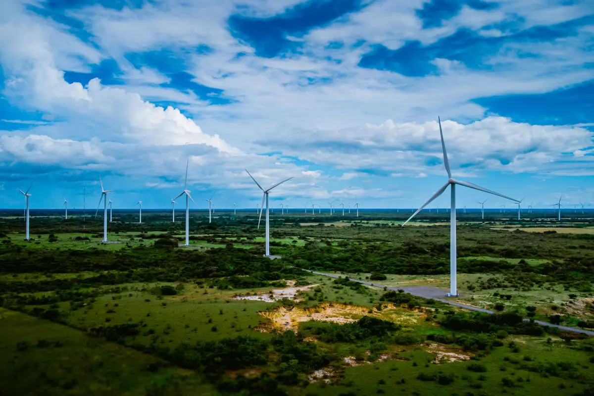 Vista aérea oblíqua de um vasto campo plano verdejante preenchido por dezenas de turbinas eólicas brancas em atividade, sob um céu azul repleto de nuvens volumosas.