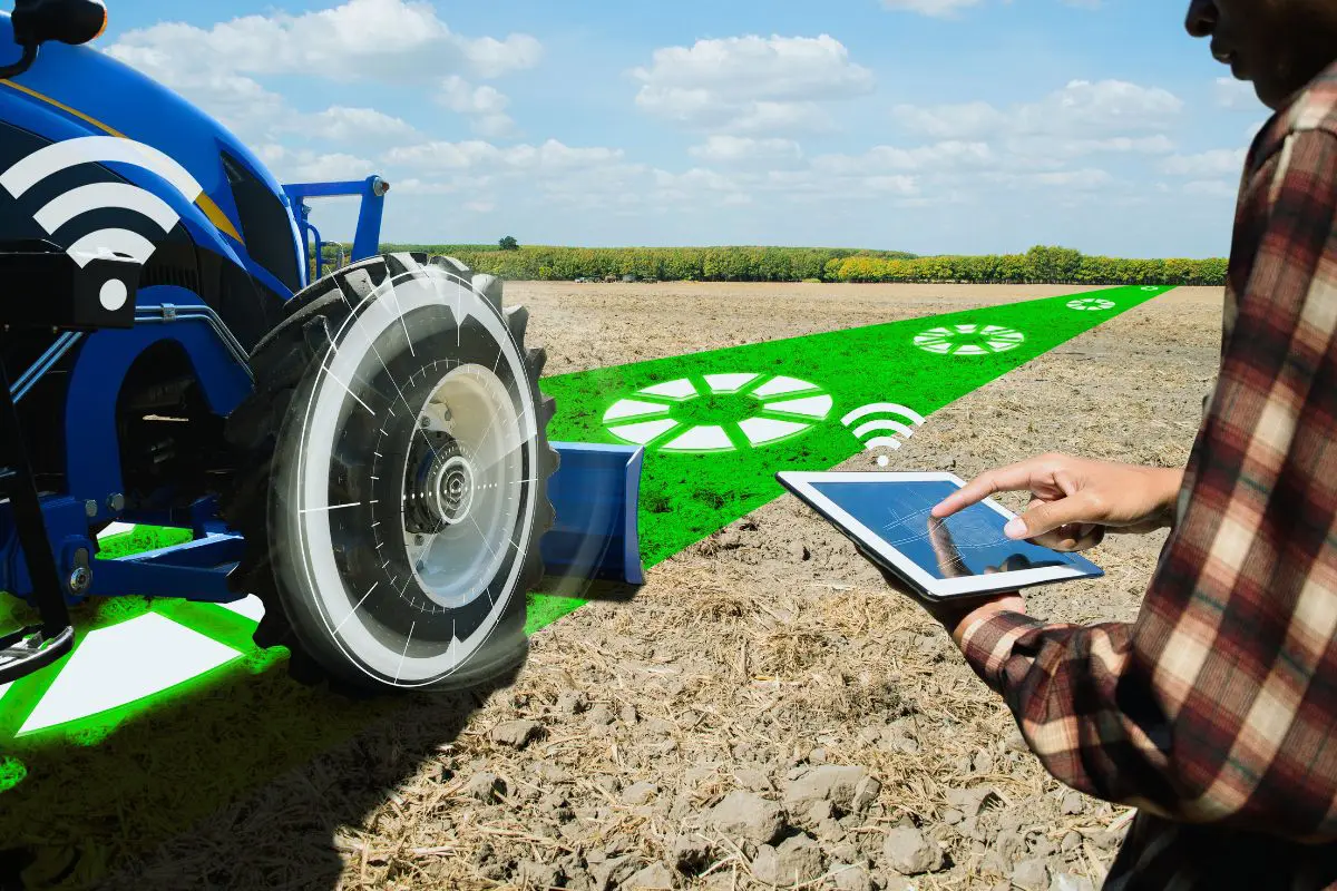 Vista parcial de um trator azul com gráficos tecnológicos no pneu e sinal de Wi-Fi, ao lado de um homem em camisa xadrez operando um tablet que projeta um caminho verde luminoso no solo arado.
