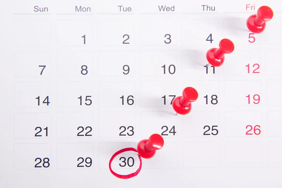 Vista superior de um calendário mensal impresso em papel branco com os números dos dias em preto e vermelho, exibindo quatro alfinetes de mapa vermelhos fincados em diagonal e o dia trinta circulado com caneta vermelha.