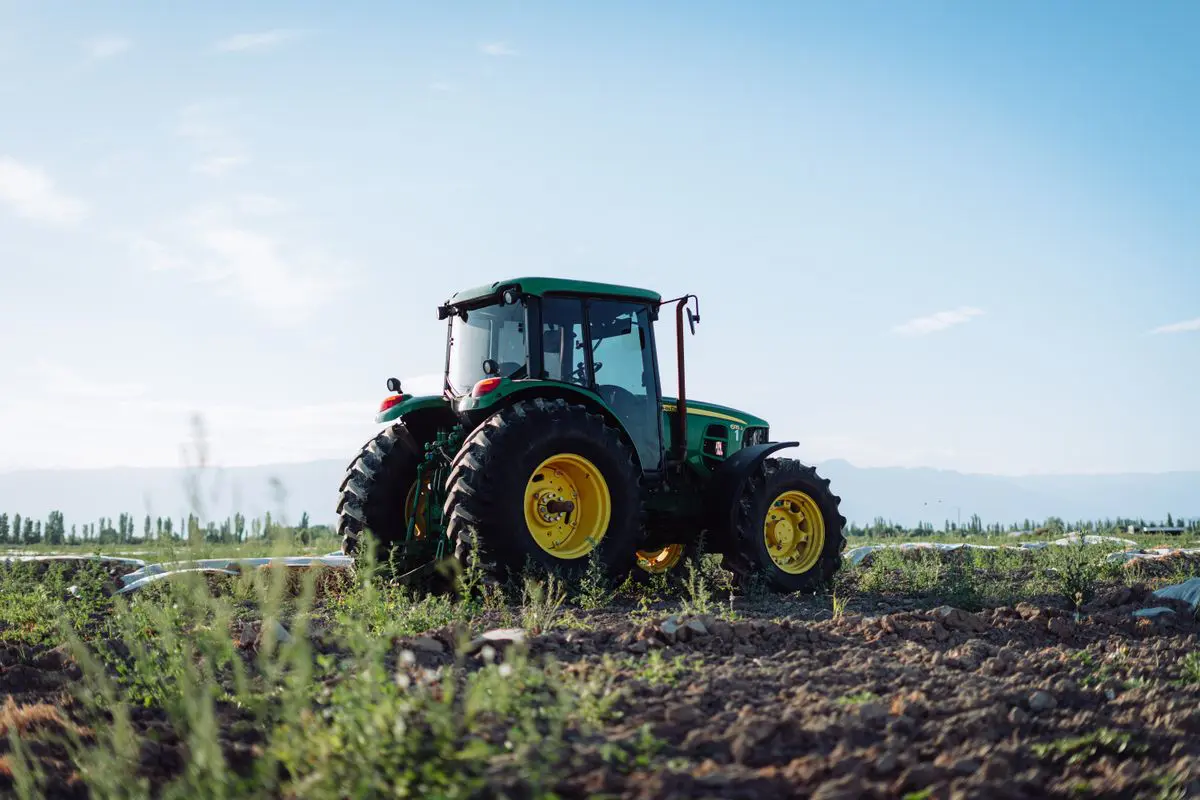 Trator agrícola verde com grandes rodas de aros amarelos posicionado em um terreno de terra arada com vegetação rasteira, sob um céu azul claro.