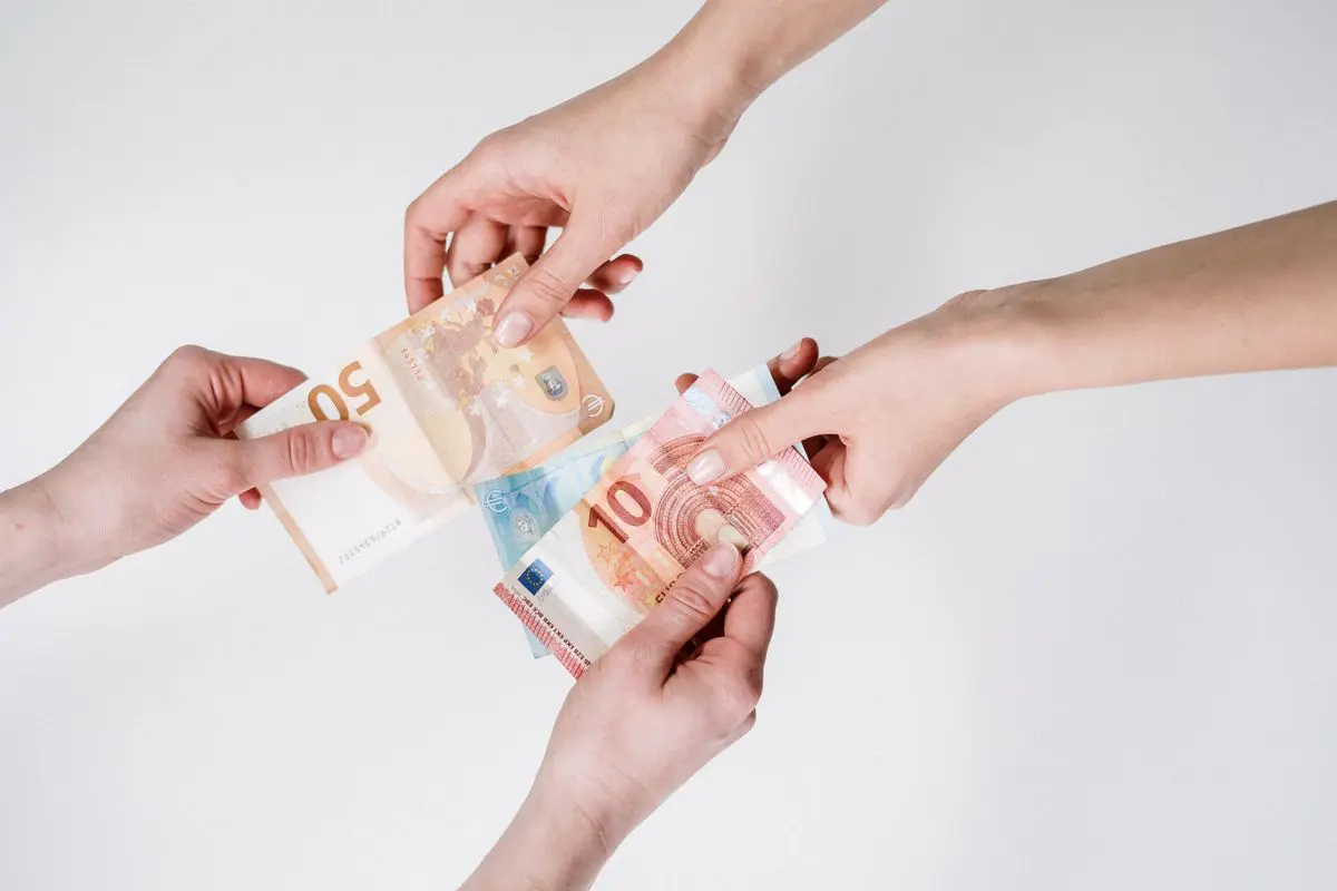 Vista superior em plano fechado de quatro mãos que se encontram ao centro sobre um fundo branco, segurando e compartilhando cédulas de Euro.