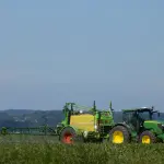 Trator agrícola verde com rodas de aros amarelos puxando um grande pulverizador acoplado em meio a uma lavoura verdejante, sob um céu azul limpo.