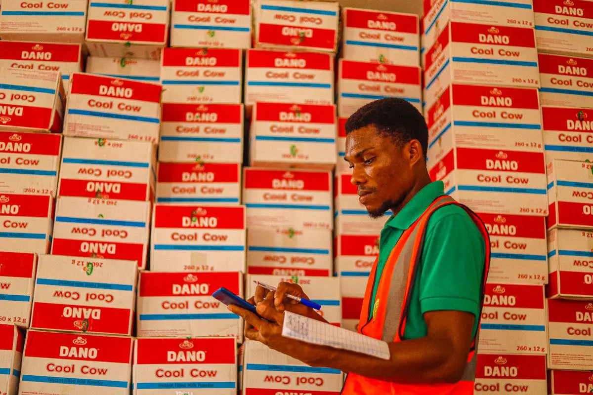 Homem jovem de pele negra vestindo camisa verde e colete refletivo laranja, de perfil, analisando um bloco de notas em um galpão repleto de caixas empilhadas da marca "DANO Cool Cow".