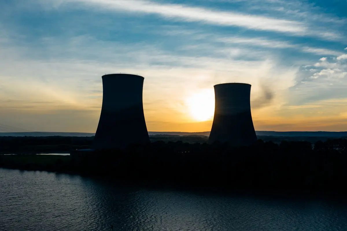 Silhueta de duas grandes torres de resfriamento hiperbólicas em contra-luz à beira de um corpo d'água ao pôr do sol, com o sol se pondo entre elas sob um céu com nuvens esparsas.