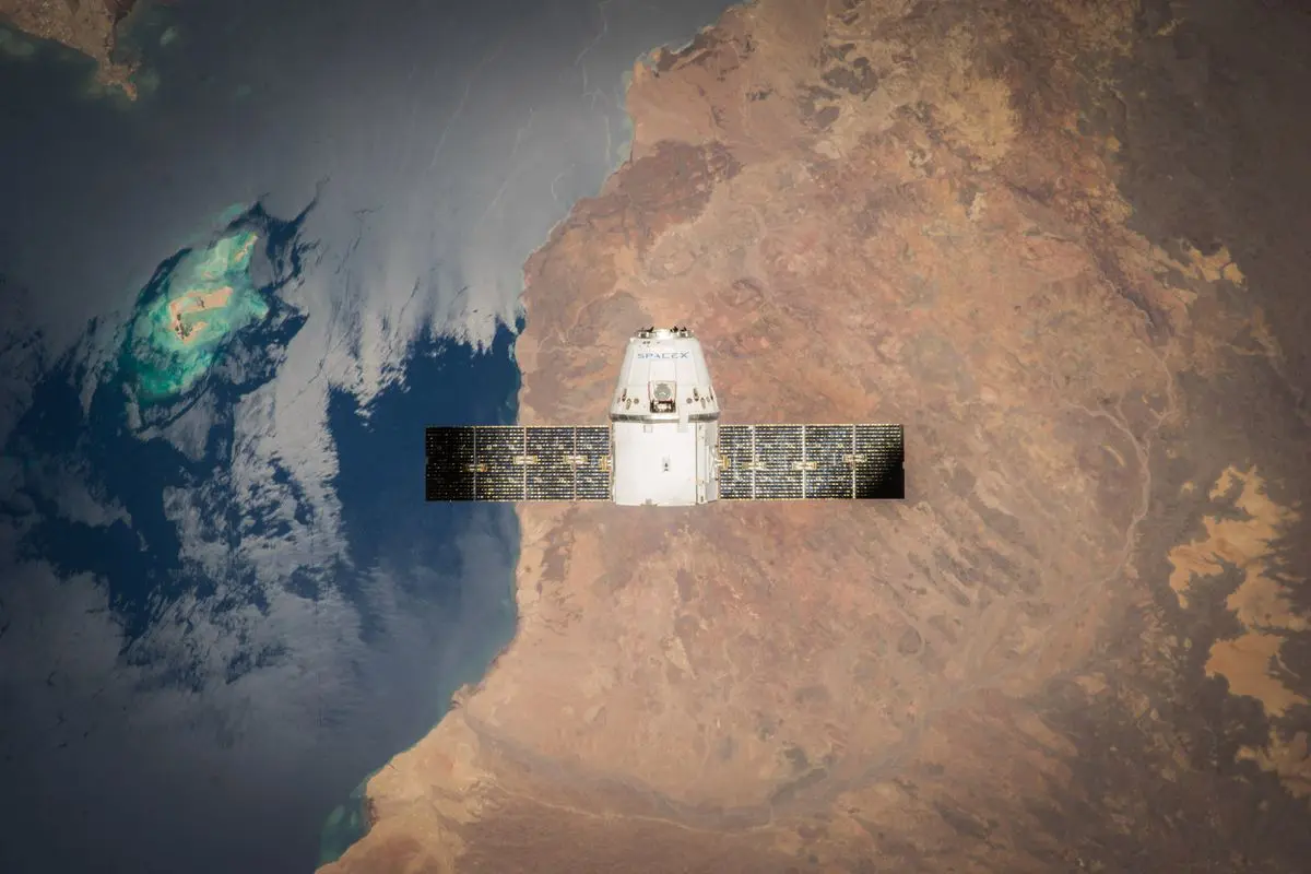 Vista aérea orbital de um satélite ou cápsula espacial branca com painéis solares horizontais estendidos, flutuando sobre uma linha de costa que divide o oceano azul escuro de uma vasta extensão de terra árida marrom.