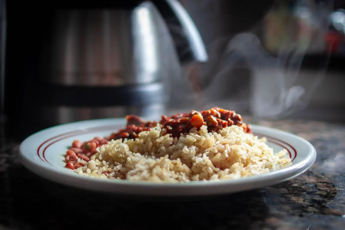 Close de um prato de comida com arroz integral, feijão e um ensopado com carne e cenoura saindo fumaça quente, sobre uma bancada de granito escura com uma cafeteira ao fundo.