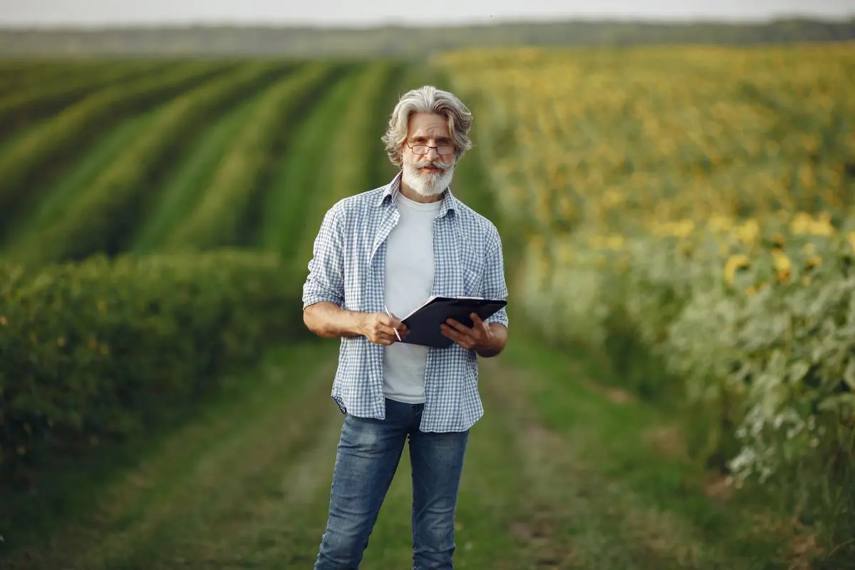 Homem de cabelos e barba grisalhos vestindo camisa xadrez e óculos, em pé no centro de um caminho entre duas plantações diferentes, segurando uma prancheta preta.