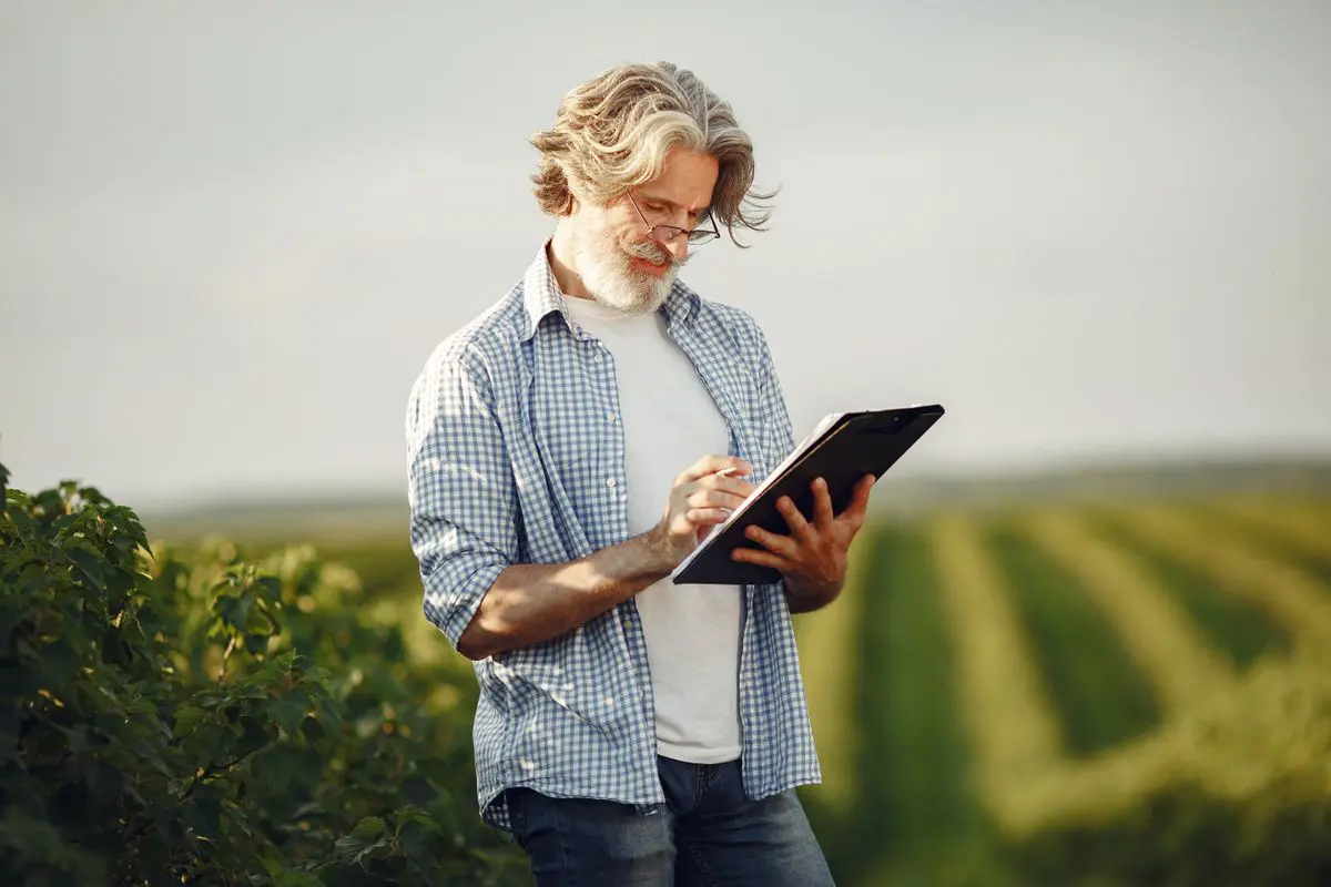 Plano médio de um homem de cabelos grisalhos e óculos, vestindo camisa xadrez azul e branca, em pé em um campo agrícola enquanto faz anotações em uma prancheta preta.