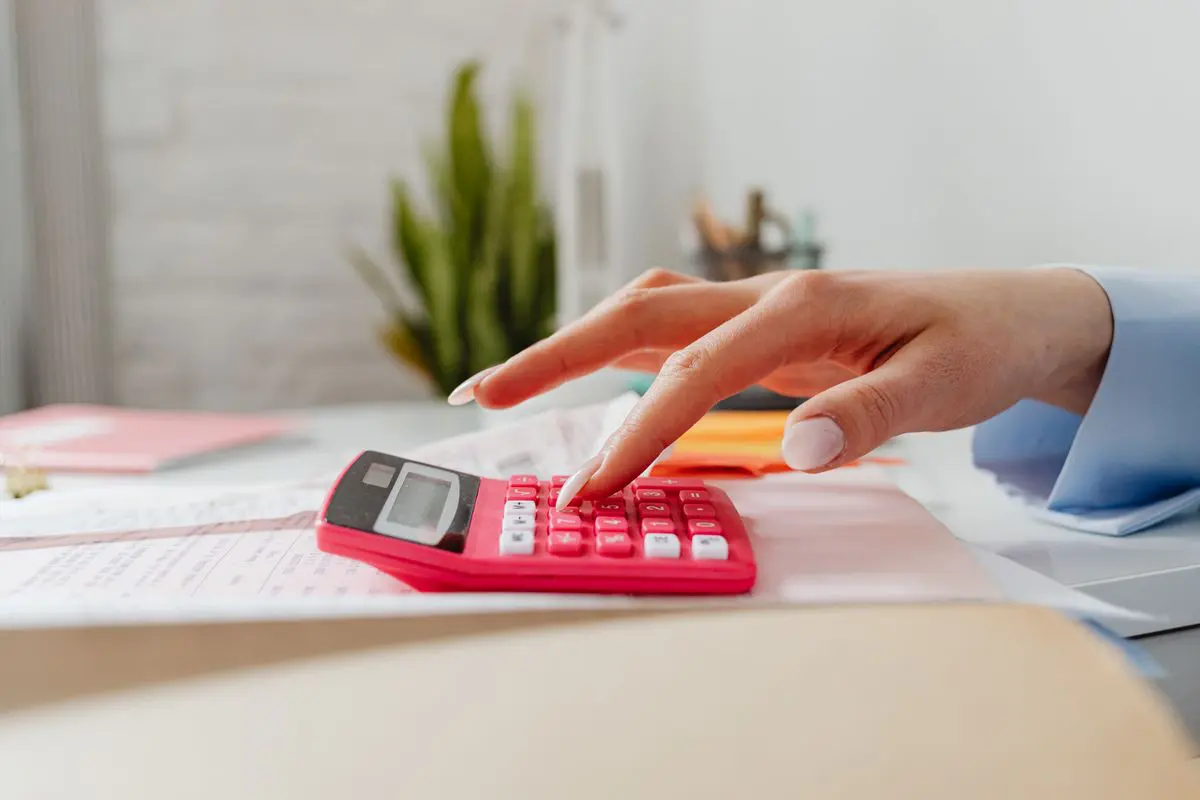 Close lateral da mão de uma pessoa digitando em uma pequena calculadora rosa sobre papéis e notas fiscais em uma mesa de escritório branca.