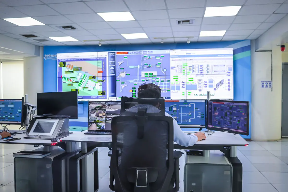 Vista traseira de um operador de costas, sentado em uma cadeira ergonômica preta em frente a múltiplos monitores de computador e a um grande painel de video wall que exibe mapas, gráficos e fluxogramas de dados em uma moderna sala de controle iluminada.