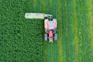 Vista aérea zenital de um grande trator vermelho acoplado a uma segadora verde cortando uma plantação de forragem intensamente verdejante.