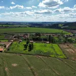 Vista aérea panorâmica de propriedades rurais com pequenas casas, galpões e estradas cercadas por vastos campos de pastagem e plantações verdes sob um céu azul com nuvens.