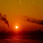 Silhueta de um complexo industrial com duas chaminés expelindo fumaça escura contra um céu de pôr do sol intensamente alaranjado e avermelhado, com o sol circular próximo ao horizonte.