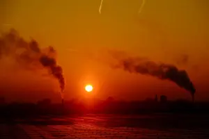Silhueta de um complexo industrial com duas chaminés expelindo fumaça escura contra um céu de pôr do sol intensamente alaranjado e avermelhado, com o sol circular próximo ao horizonte.