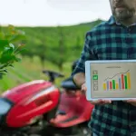 Homem com camisa xadrez segurando um tablet que exibe um gráfico de barras colorido intitulado "HARVEST LEVEL", em frente a uma plantação verde e um pequeno trator vermelho desfocados.