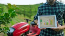 Homem com camisa xadrez segurando um tablet que exibe um gráfico de barras colorido intitulado "HARVEST LEVEL", em frente a uma plantação verde e um pequeno trator vermelho desfocados.