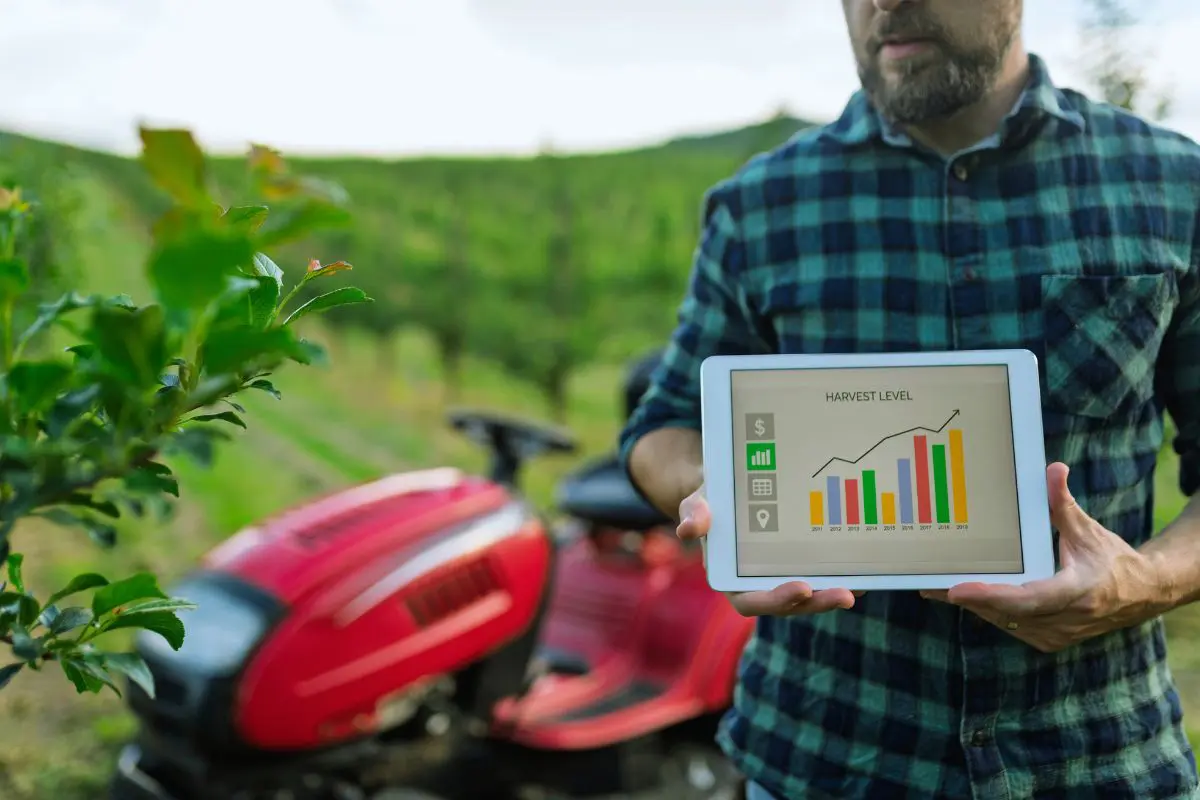 Homem com camisa xadrez segurando um tablet que exibe um gráfico de barras colorido intitulado "HARVEST LEVEL", em frente a uma plantação verde e um pequeno trator vermelho desfocados.