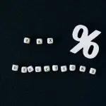 Pequenos cubos brancos com letras pretas formam as palavras "TAX" e "DEDUCTIONS" sobre um fundo preto texturizado, ao lado de um grande símbolo de porcentagem branco.