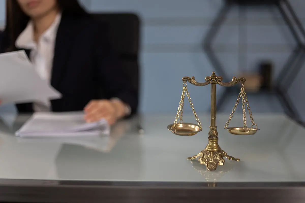 Plano médio de uma balança da justiça de metal dourado em primeiro plano sobre uma mesa de vidro, com uma mulher de terno preto folheando papéis desfocada ao fundo.